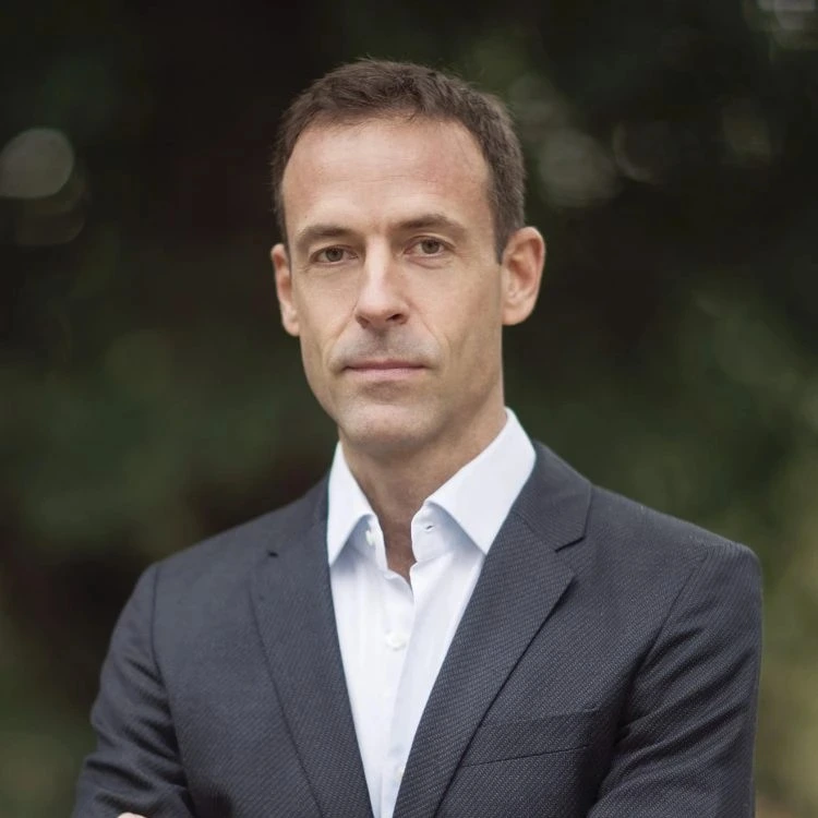 Dr. Robert Morrison in a dark suit with a white shirt stands outdoors with arms crossed, looking confidently at the camera.