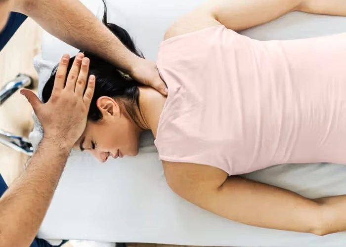 A person in a pink sleeveless shirt lies face down on a table, receiving a neck massage to relieve a stiff neck.