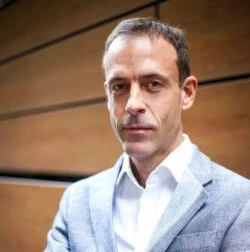 Dr. Robert Morrison in a light gray suit with a white shirt stands confidently with arms crossed, posing in front of a wooden panel wall.